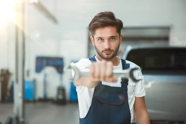 Joven mecánico guapo vistiendo uniforme celebración llave en su mano en coche centro de servicio — Foto de Stock