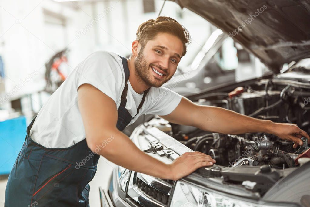 young smiling handsome mechanic in uniform fixing motor in car bonnet working in service center looking happy