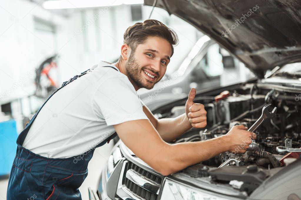 young smiling mechanic in uniform fixing motor problems in car bonnet working in service center showing like sign