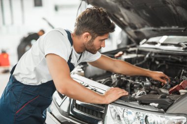 Üniformalı genç yakışıklı tamirci araba kaputunda motor tamir ediyor servis merkezinde çalışıyor.