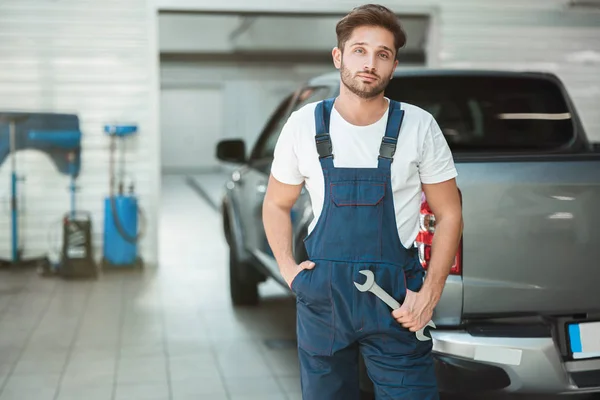 Joven mecánico guapo vistiendo uniforme con una mano en el bolsillo y con llave en otra mano se levanta en el centro de servicio del coche — Foto de Stock