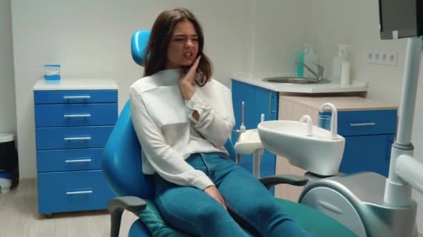 Молодая красивая брюнетка пациентка чувствует зубную боль касается ее щеки, сидя в кабинете стоматолога — стоковое видео