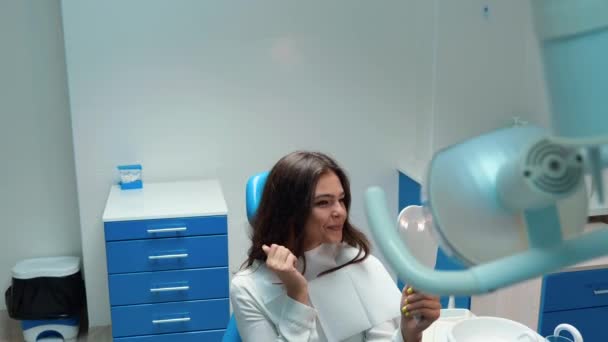 若い美しいブルネットの女性患者は歯科医のオフィスに座っている間に鏡を見て結果に満足そうに見えます — ストック動画