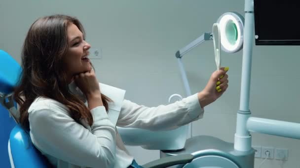 Μελαχρινή γυναίκα ασθενής φαίνεται ευχαριστημένος με αποτέλεσμα βλέποντας στον καθρέφτη γελώντας και χαμογελώντας, ενώ κάθεται στο γραφείο οδοντίατροι — Αρχείο Βίντεο