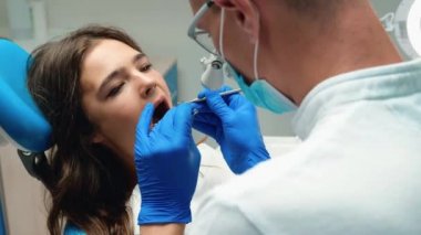 Genç dişçi, tıbbi eldiven ve maske takıyor. Gözlük takıyor. Esmer kadınların dişlerini kontrol ediyor.
