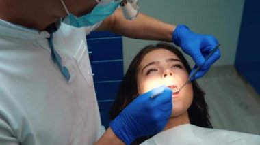 Tıbbi eldiven, maske ve gözlük takan genç dişçi, dişçi muayenehanesinde diş aynası ve baraj kullanarak kadınların dişlerini iyileştirir.