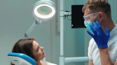 Tıbbi eldiven, maske ve gözlük takmış genç yakışıklı dişçi dişçi dişçi muayenesinden önce dişçi muayenesinden önce güzel bir kadın hastayla tartışıyor.