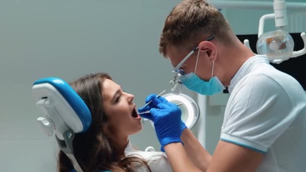 Молодой стоматолог в медицинских перчатках и маске проверки молодых брюнеток пациентки проблемы с зубами с помощью стоматологических инструментов — стоковое видео
