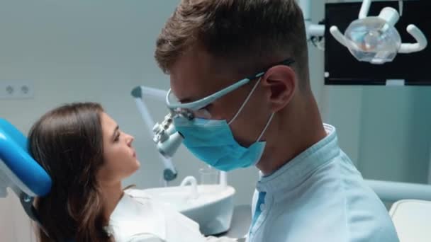 Молодой стоматолог в медицинских перчатках и маске проверки молодых брюнеток пациентки зуб пациента с помощью стоматологических инструментов пациент выглядит счастливым — стоковое видео