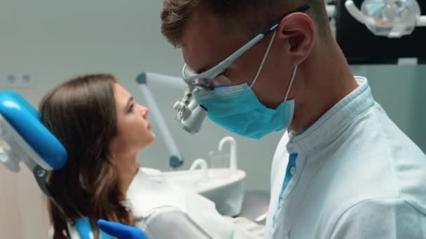 Ung tandläkare i medicinsk mask justerar blå handskar gör sig redo att börja undersöka unga brunett kvinnor patient — Stockvideo