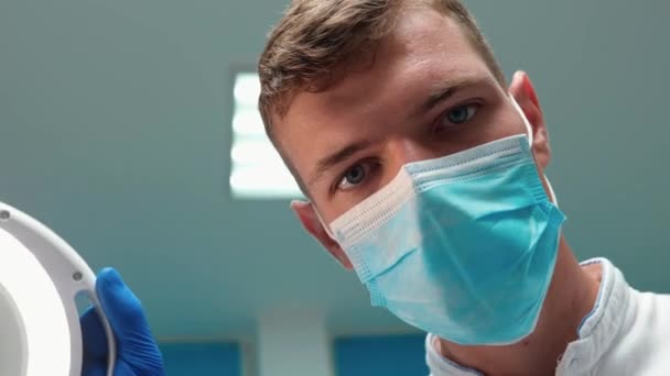 Jonge knappe arts tandarts in masker houdt medische lamp tijdens het onderzoek van patiënten en beweegt dichter — Stockvideo
