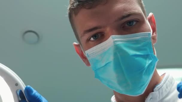 Jeune beau médecin dentiste en masque tient lampe médicale pendant l'examen des patients à l'aide d'un miroir dentaire — Video