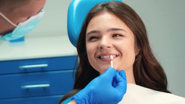 Lächelnd brünett Patientin Frau mit Termin in Zahnklinik Zahnarzt in Maske und Handschuhen gilt Emaille-Skala, um den richtigen Schatten zu holen — Stockvideo