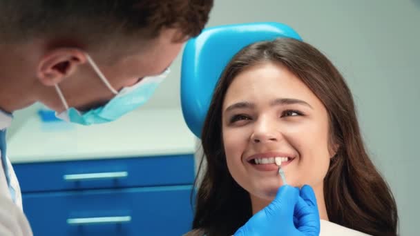 マスクと手袋の若い歯科医は、歯科クリニックでの任命中に患者の女性を笑顔ブルネットのための右の日陰を拾うためにエナメルスケールを適用します — ストック動画