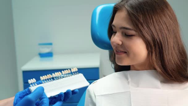 Стоматолог в перчатках демонстрирует эмалевую шкалу, чтобы подобрать правильный оттенок для улыбающейся брюнетки пациентки во время приема в стоматологической клинике — стоковое видео