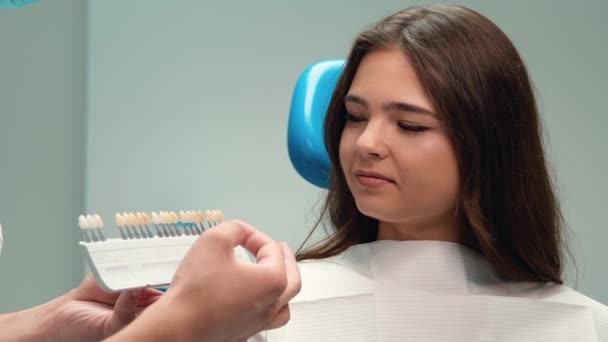 Dentysta pokazuje skalę emalii, aby podnieść odpowiedni odcień do brunetka pacjenta kobieta podczas wizyty w klinice stomatologicznej — Wideo stockowe