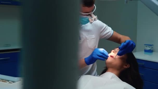 Jovem dentista vestindo luvas médicas, máscara e óculos sela os dentes das mulheres pacientes usando espelho dental e represa durante consulta na clínica odontológica — Vídeo de Stock