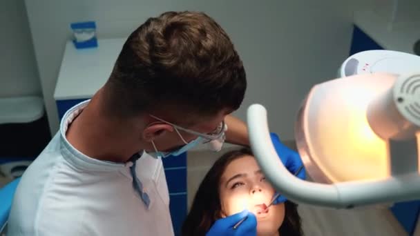 Tıbbi eldiven, maske ve gözlük takan genç dişçi, dişçi muayenehanesinde diş aynası ve baraj kullanarak kadın dişlerini kontrol ediyor ve tedavi ediyor. — Stok video