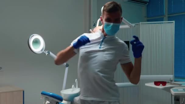 Funny young handsome doctor dentist in medical gloves and mask dancing happily near his working place in his office — Stock Video