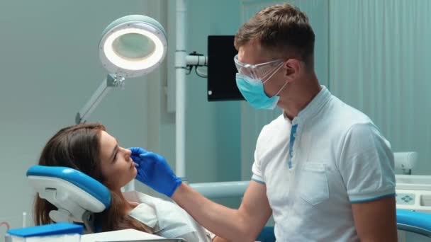 年轻英俊的牙医，戴着医用蓝色手套、面具和眼镜，在牙科诊所检查前与美丽的病人讨论牙科治疗过程 — 图库视频影像