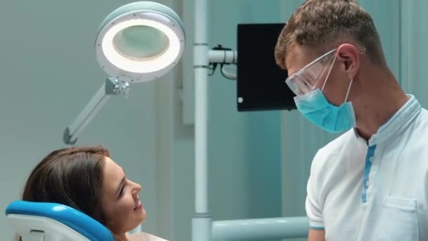 Ung stilig tandläkare i medicinsk blå handskar, mask och glasögon diskuterar med brunett patient kvinna process tandvård innan undersökning på tandvårdskliniken — Stockvideo