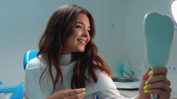 Young beautiful brunette woman patient looks pleased with result watching in the mirror touching her teeth smiling while sitting in the dentists office — Stock Video