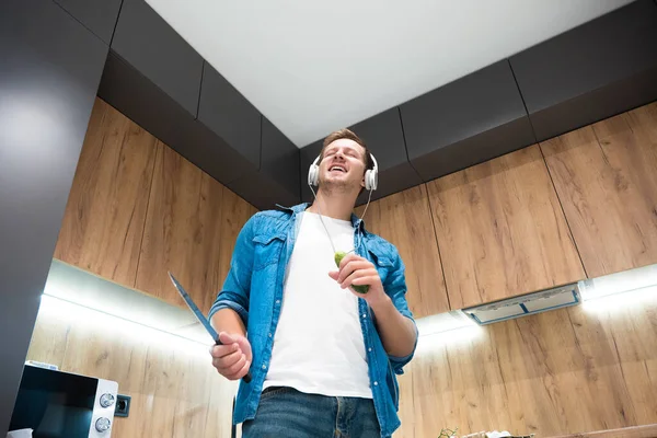 Joven guapo hombre en auriculares bailando en la cocina con cuchillo en una mano y pepino en otra — Foto de Stock