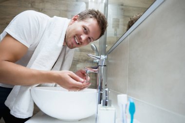 Genç, yakışıklı, gülümseyen adam yüzünü banyoda suyla yıkar.
