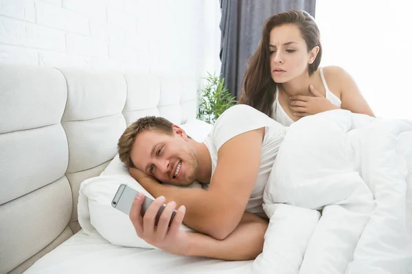 Przystojny uśmiechnięty mężczyzna z telefon komórkowy leżący w łóżku z młoda piękna kobieta zły na niego — Zdjęcie stockowe