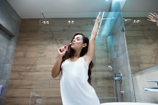 Jong mooi glimlachen vrouw in wit toga houdt tandenborstel als een microfoon en zingt in de badkamer terwijl het doen van haar ochtend routine — Stockfoto