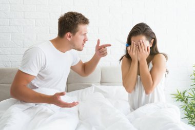 Genç bir çift, kavga eden yakışıklı bir adamı, yatakta oturmuş elleriyle yüzünü kapatan üzgün bir kadını işaret ediyor.