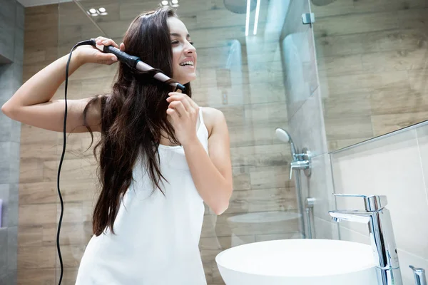 Giovane bella donna sorridente in abito bianco in piedi in bagno arricciando i capelli lunghi bruna con il ferro da stiro — Foto Stock