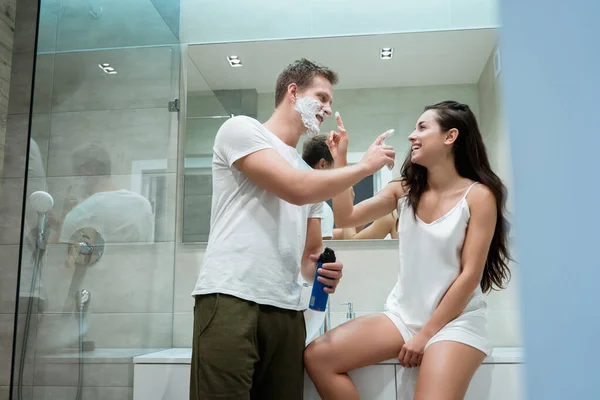 Joven hombre guapo y hermosa mujer divirtiéndose en el baño mientras se aplica espuma de afeitar y pone un poco en su cara —  Fotos de Stock
