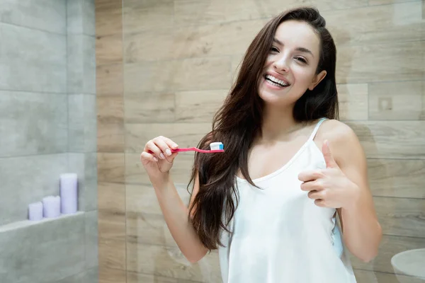 Joven hermosa mujer de pie delante del espejo del baño mientras se cepilla los dientes mostrando como signo —  Fotos de Stock