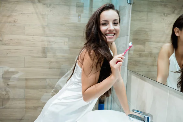 Joven hermosa mujer sonriente de pie junto al espejo del baño mientras se cepilla los dientes —  Fotos de Stock