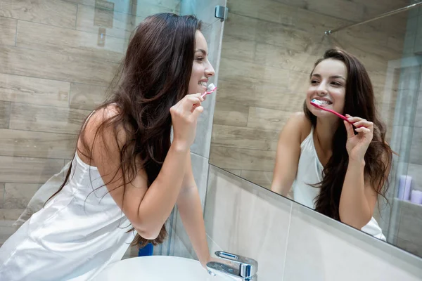 Giovane bella donna sorridente in asciugamano bianco in piedi accanto allo specchio del bagno mentre si lava i denti — Foto Stock