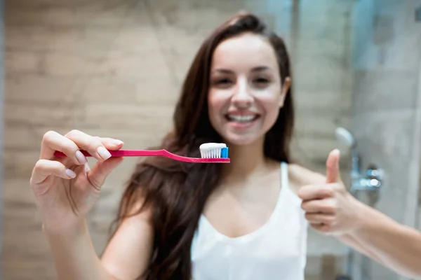 Joven hermosa mujer sonriente sostiene cepillo de dientes con pasta de dientes en el baño mostrando como signo —  Fotos de Stock