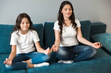 Genç, güzel anne ve genç kızı oturma odasındaki kanepede yoga pozisyonunda oturuyorlar.