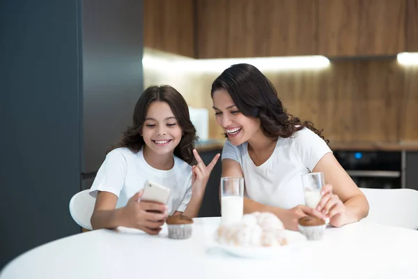 Joven madre con su hija adolescente viendo fotos en el teléfono mientras toma cupcakes con leche para el desayuno ambos sonriendo — Foto de Stock