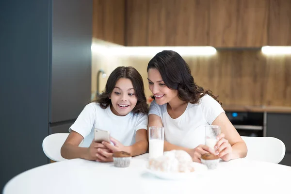 Mooie jonge moeder met haar schattige tiener dochter kijken iets grappigs aan de telefoon terwijl het hebben van cupcakes met melk voor het ontbijt zowel glimlachen — Stockfoto