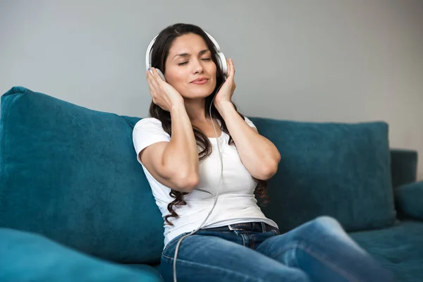 Joven hermosa mujer en auriculares escuchando su música favorita en la sala de estar sentado en el sofá — Foto de Stock