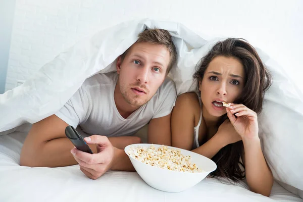 Young couple handsome man and beautiful woman watching TV in bed with pop corn looking surprised — Stock Photo, Image