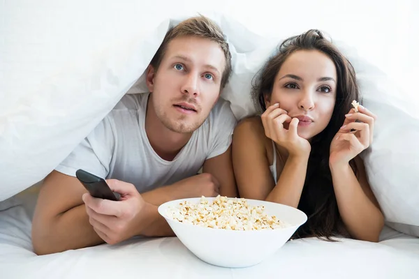 Young couple handsome man and beautiful woman watching TV in bed eating pop corn looking fascinated — Stock Photo, Image