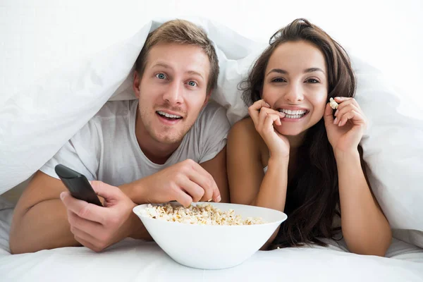 Young couple handsome man and beautiful woman watching TV in bed eating pop corn looking happy — Stock Photo, Image