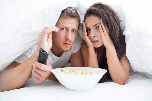 Young couple handsome man and beautiful woman watching horror movie in bed eating pop corn looking frightened — Stock Photo, Image