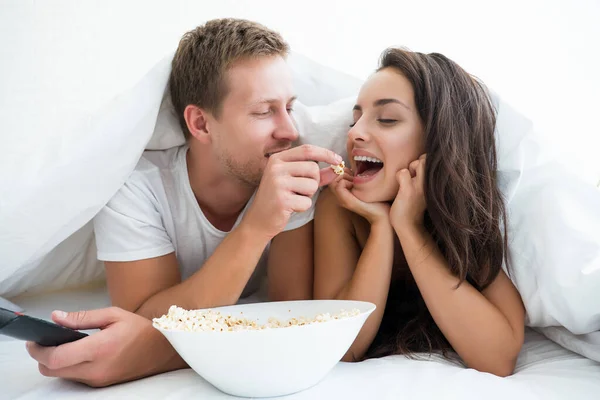 Young couple handsome man feeding beautiful woman popcorn with his hand watching movie in bed looking happy — Stock Photo, Image