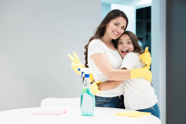 Joven hermosa mujer abrazando a su linda hija adolescente en guantes amarillos haciendo limpieza niño muestra signo de victoria — Foto de Stock