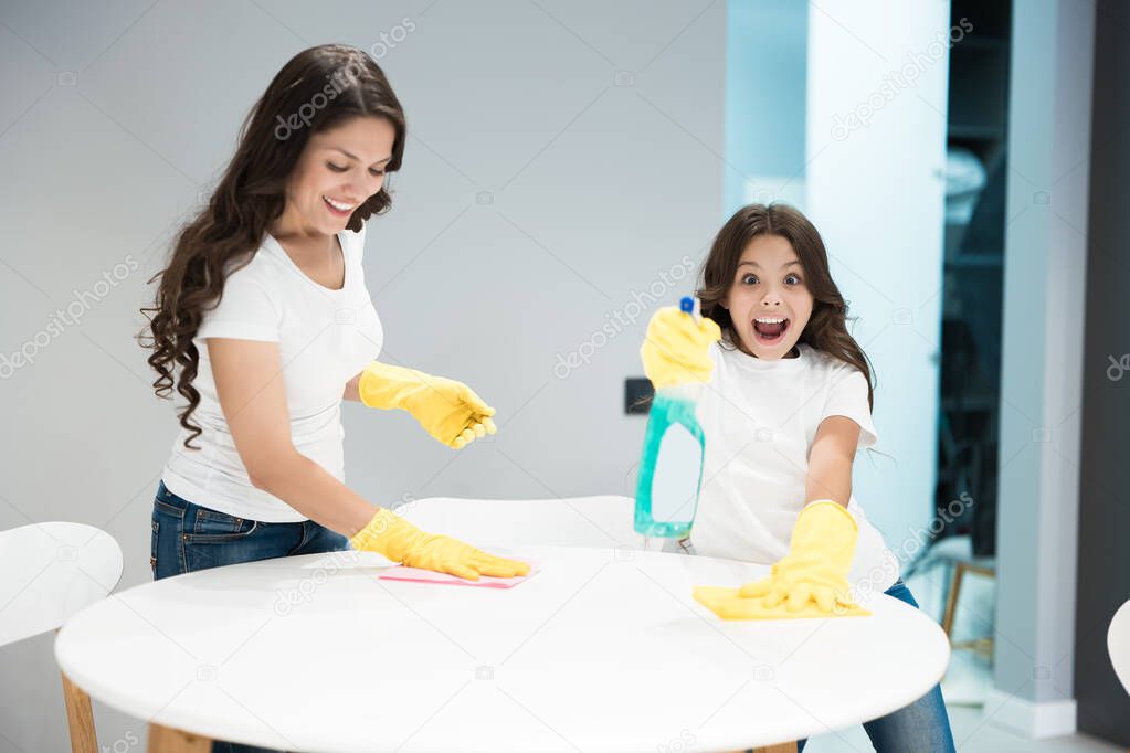 young beautiful woman laughing while her cute teen daughter spraying detergent to wipe dust off the table while cleaning the house