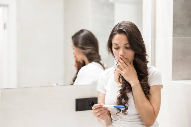 Genç, güzel, şaşkın bir kadın tuvalette oturuyor ve hamilelik testi yapıyor.