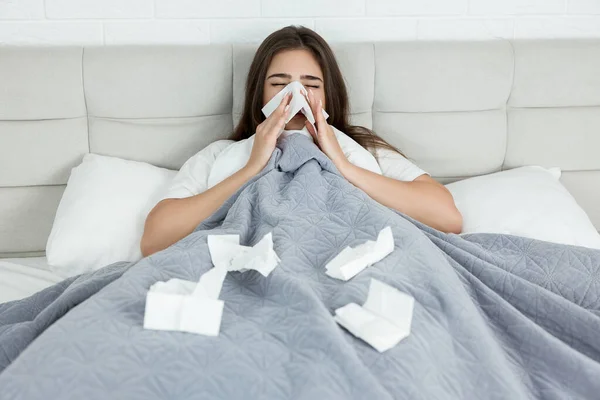 young sick woman sitting in bed bowing out her running nose with paper towel early in the morning